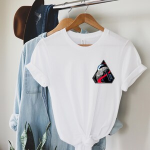 May include: White t-shirt with a triangular graphic featuring a moon, a red streak, and the text "III". The shirt is hanging on a wooden hanger with a blue denim shirt and a brown hat in the background.