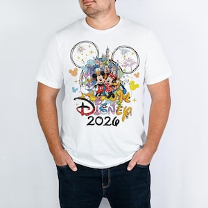May include: White t-shirt featuring a colorful graphic of Disney characters, a castle, fireworks, and the text "Disney 2026." The design includes Mickey and Minnie Mouse, Donald Duck, and Goofy. The shirt has a crew neck and short sleeves.