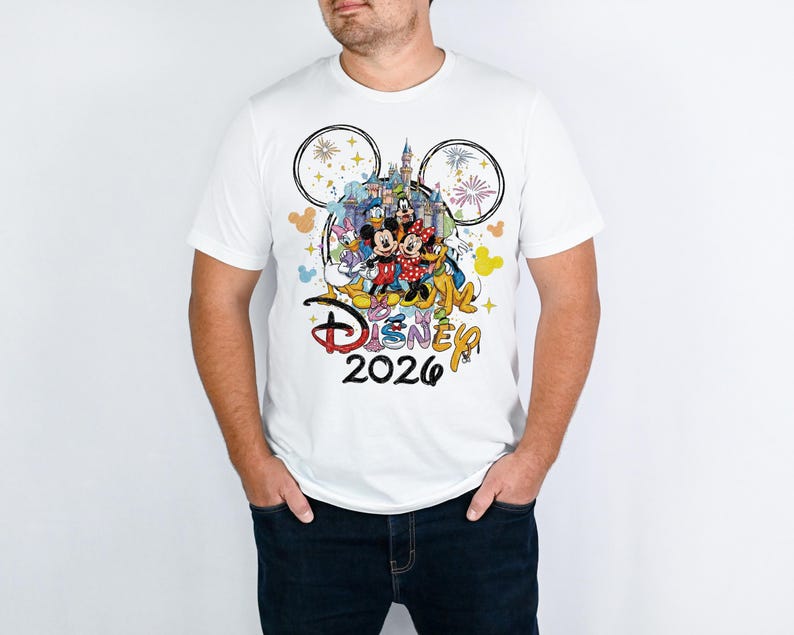 May include: White t-shirt featuring a colorful graphic of Disney characters, a castle, fireworks, and the text "Disney 2026." The design includes Mickey and Minnie Mouse, Donald Duck, and Goofy. The shirt has a crew neck and short sleeves.