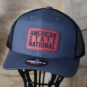 May include: A gray trucker hat with a black mesh back and a curved brim. The front features a red rectangular patch with the words "AMERICAN STATE NATIONAL" in white block letters. The hat is displayed on a black metal stand.