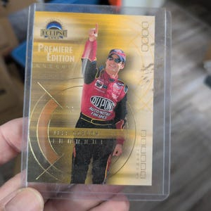 May include: A 2002 Eclipse Premiere Edition trading card featuring Jeff Gordon in a red and black racing suit with DuPont branding. The card has a gold background with circular graphic elements, the text Eclipse 2002, and Premiere Edition.