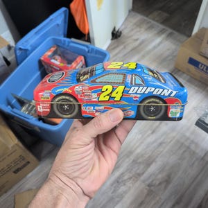 May include: A blue and red metal tin shaped like a NASCAR race car featuring the number 24, DuPont branding, and various sponsor logos including Pepsi, Goodyear, and Fritos. The car has a flame design on the hood and side panels.