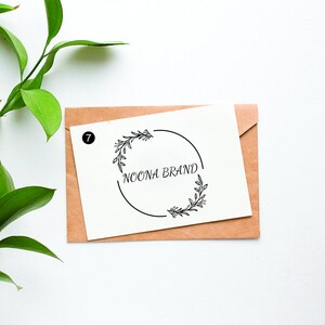 Puede incluir: Una tarjeta blanca con un sobre marrón detrás. La tarjeta tiene una corona de flores en blanco y negro con el texto "NOONA BRAND" dentro de la corona.
