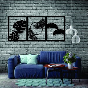 Pode incluir: Três peças de decoração de parede de metal preto com designs de folhas tropicais recortadas. As peças estão dispostas em uma fila em uma parede de tijolo. Um sofá azul com almofadas está em primeiro plano.