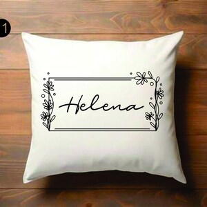 Puede incluir: Cojín cuadrado blanco con el nombre "Helena" en escritura cursiva, enmarcado por un borde rectangular negro con detalles florales. El fondo es de madera.