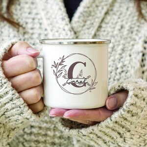 Könnte beinhalten: Weiße Emailtasse mit braunem Blumendesign und dem Buchstaben "C" in einem Kreis. Der Name "Carol" steht unter dem Buchstaben.