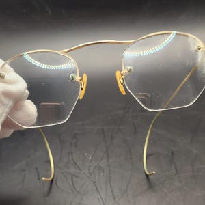May include: Rimless eyeglasses featuring a thin gold-tone metal frame with a high bridge and curved temple arms. The clear lenses have an octagonal shape and are secured to the frame with small screws at the outer edges and nose bridge area.