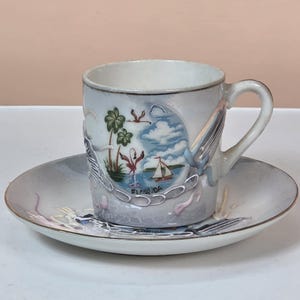 May include: A vintage teacup and saucer set. The cup and saucer are white with a decorative design featuring a Florida landscape with palm trees, a sailboat, and the word "Florida". The set has a gold trim.