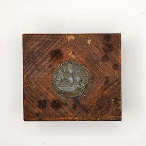 May include: A wooden box with a diamond pattern on the lid. The lid has a metal inlay of a ship with sails.