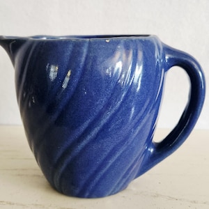 May include: A cobalt blue ceramic pitcher with a handle and a spout. The pitcher has a textured, swirled design. The handle is a curved, open loop. The pitcher is likely vintage and suitable for serving beverages.