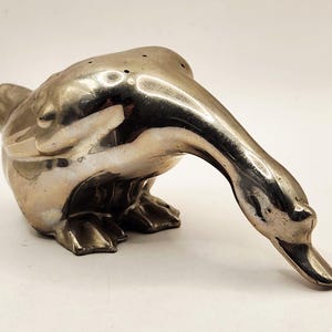 May include: A polished silver-toned metal goose figurine shaped as a shaker. The goose is depicted in a forward-leaning posture with its head bowed downward. The top of the goose's back features a series of small, evenly spaced holes for dispensing.