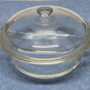 May include: A clear glass casserole dish with a matching lid. The dish has two handles and a round knob on the lid. The glass is transparent, allowing a view of the contents. The dish is likely used for baking or serving food.