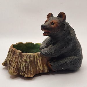 May include: A ceramic bear figurine with a dark brown body and a lighter brown face and ears. The bear is seated next to a tree stump-shaped planter with a green interior. The planter has a textured, wood-like appearance.