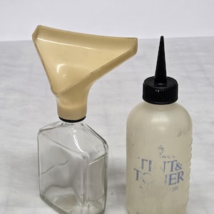 May include: Two bottles, one clear glass with a cream-colored funnel, and a translucent plastic bottle with a black cap. The plastic bottle has the text "TINT & TONER" printed on it. Both bottles are on a white surface.