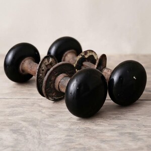 May include: Two vintage door knobs featuring smooth, round, black heads attached to weathered, rusted metal spindles and circular backplates. The hardware displays signs of age with oxidized metal surfaces and worn, dark, glossy spherical handles.