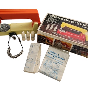 May include: An orange and cream-colored rhinestone and stud setter machine with accessories, a box, and instruction booklets. The box reads "The Rhinestone & Stud Setter by Ronco" and "As seen on TV". A silver-toned necklace is also included.