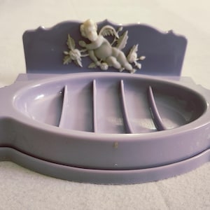 May include: A lavender-colored soap dish with a decorative cherub and floral relief. The dish has a scalloped back and a slotted base. The cherub is white, and the dish is made of a smooth, solid material. A vintage bathroom accessory.