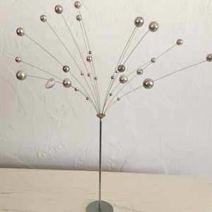 May include: A silver metal sculpture with a central stem and radiating wires. Each wire has a silver sphere at the end, creating a starburst effect. The sculpture is on a round base and sits on a light-colored surface.