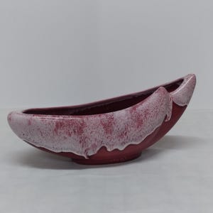 May include: A pink and white ceramic planter shaped like a canoe. The planter has a speckled glaze and a white rim.