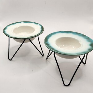 May include: Two ceramic bowls with a white interior and a teal and white rim. The bowls are supported by black metal stands. The stands have three legs that meet in the center. The bowls have a unique, hat-like shape.