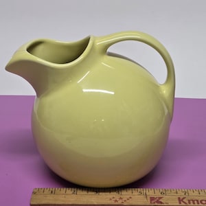 May include: A pale yellow ceramic pitcher with a rounded body and a curved handle. The pitcher has a spout for pouring and is likely used for serving beverages. The pitcher is sitting on a purple surface.