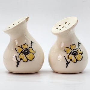 May include: A pair of white ceramic salt and pepper shakers with a floral design. The shakers have a yellow flower with black outlines on a white background.
