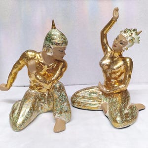 May include: Two ceramic figurines depicting traditional dancers. The figures are kneeling with arms raised, and are painted in gold with green and white accents. The figurines have detailed facial features and are wearing ornate headwear.