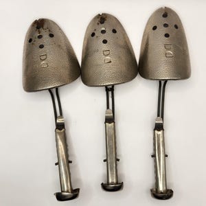 May include: Three vintage metal shoe trees. Each tree has a textured, silver-toned toe piece with small holes and the letters "DD". The trees have a black wire and a silver-toned adjustable handle.