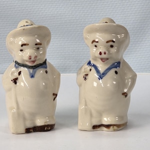 May include: A pair of ceramic salt and pepper shakers shaped like pigs. Each pig wears a hat, a blue collar, and has brown details. The shakers are off-white with a glossy finish.