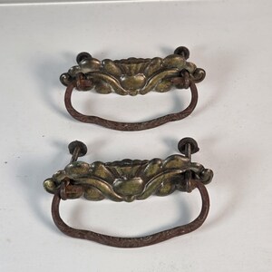 May include: Two matching drawer pulls featuring a decorative brass-toned backplate with a scalloped edge and a central embossed design. Each pull includes a dark, textured, U-shaped metal drop handle attached to the backplate with two threaded bolts.