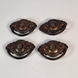 May include: A set of four oval-shaped metal drawer pulls with a dark bronze finish. Each pull features an ornate, embossed border, a central circular plate with a small decorative knob, and a hinged, semi-circular drop handle for gripping.