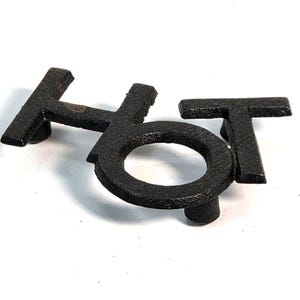 May include: A black metal trivet in the shape of the word "HOT". The letters are in a bold, blocky font and have a textured surface. The trivet is designed to protect surfaces from hot items.
