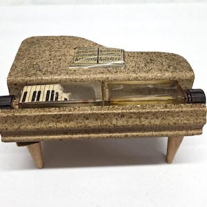 May include: A miniature piano-shaped perfume bottle with a textured, speckled beige finish. The piano has a black and white keyboard, a music sheet, and two clear glass perfume bottles. The piano rests on four small wooden legs.