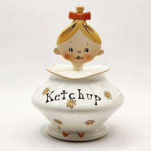 May include: A vintage white ceramic ketchup dispenser with a cartoon girl figure on top. The dispenser is decorated with small orange flowers and the word "Ketchup" in black script. The girl has blonde hair and a red bow.