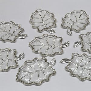 May include: Seven clear glass leaf-shaped dishes with silver-colored trim. The dishes have a detailed vein design and a small stem. The leaves are arranged on a white surface.
