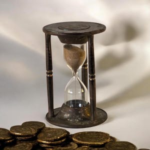 May include: An antique hourglass with a dark brown metal frame and a clear glass center. The hourglass is filled with sand, and sits on a base with a pile of gold coins in the foreground. The hourglass is a symbol of time and value.