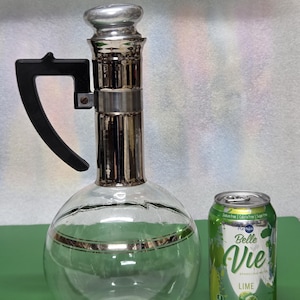 May include: A clear glass decanter with a silver-colored top and accents, a black handle, and a silver band around the middle. Next to it is a green and white can of Belle Vie Lime sparkling water.