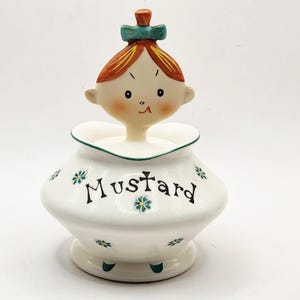 May include: A vintage ceramic mustard jar with a whimsical design. The white jar features the word "Mustard" in black script, accented with small blue flowers. The lid is a stylized head with orange hair, a green bow, and a small orange top.