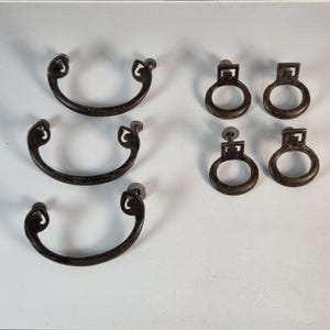 May include: A collection of seven dark metal furniture hardware pieces. Three are curved, semi-circular bail pulls with decorative scrolled ends. Four are circular ring pulls, each featuring a square mounting plate with a geometric cutout design.