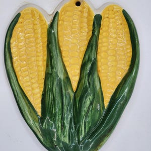 May include: A ceramic wall decor in the shape of three ears of corn. The corn is yellow with green husks. The decor has a hole at the top for hanging.