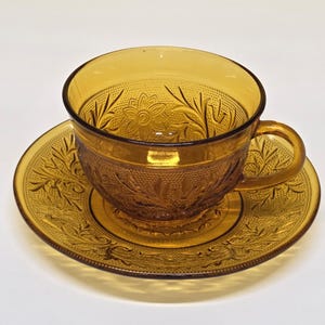 May include: Amber-colored glass teacup and saucer set. The cup and saucer feature an embossed floral and leaf design. The cup has a handle and sits on a matching saucer. The set is made of transparent glass.