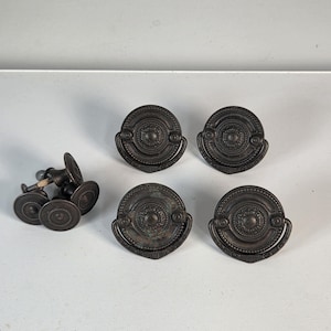 May include: A set of six circular dark bronze metal drawer pulls. Each pull features a central medallion with concentric rings and a beaded border, accompanied by a hinged drop handle. The hardware includes threaded metal posts for mounting.