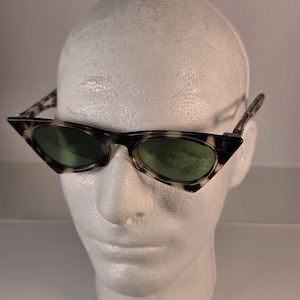 May include: Cat-eye sunglasses with a tortoiseshell pattern frame and green lenses. The sunglasses are displayed on a white foam head. The frame has a pointed, angular shape.