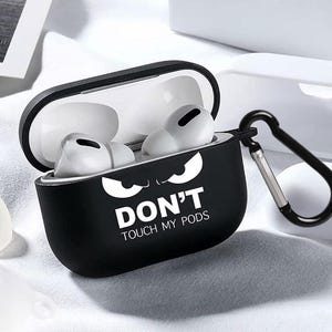 Puede incluir: Funda protectora negra para auriculares inalámbricos con un gráfico blanco de ojos enojados y el texto "DON'T TOUCH MY PODS". La funda tiene un mosquetón negro.