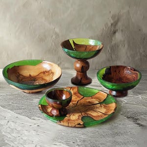 Puede incluir: Un conjunto de cuatro recipientes de madera con vetas naturales y detalles en resina verde translúcida. El set incluye un cuenco grande poco profundo, un cuenco alto con pedestal, un cuenco con pedestal más pequeño y una taza sobre un plato circular.