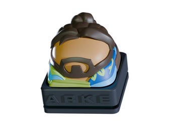May include: A custom keycap featuring a stylized character head with brown hair, a tan face, and a dark beard. The keycap has a blue and green design around the face and is set on a black base with the word "ARKE" embossed on the front.