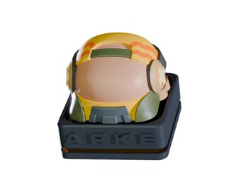 May include: A stylized, yellow and green helmet with orange accents, set in a dark gray base. The base features the letters "ABKE" in raised lettering. The helmet has a tan face shield and ear pieces.