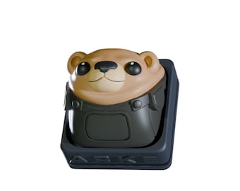 May include: A brown and black artisan keycap featuring a cartoon otter design. The otter has large black eyes and is wearing a black outfit. The keycap is set in a dark gray base with the word "ARKE" on the front.