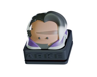 May include: A stylized, cartoon-like character keycap in shades of gray, purple, and white. The keycap sits on a dark gray base with the letters "ABKE" embossed on the front. The character has a distinctive hairstyle and facial features.