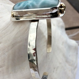 May include: A silver cuff bracelet featuring a light blue and white gemstone set in a silver bezel. The bracelet has a hammered finish and is accented with small gold beads.